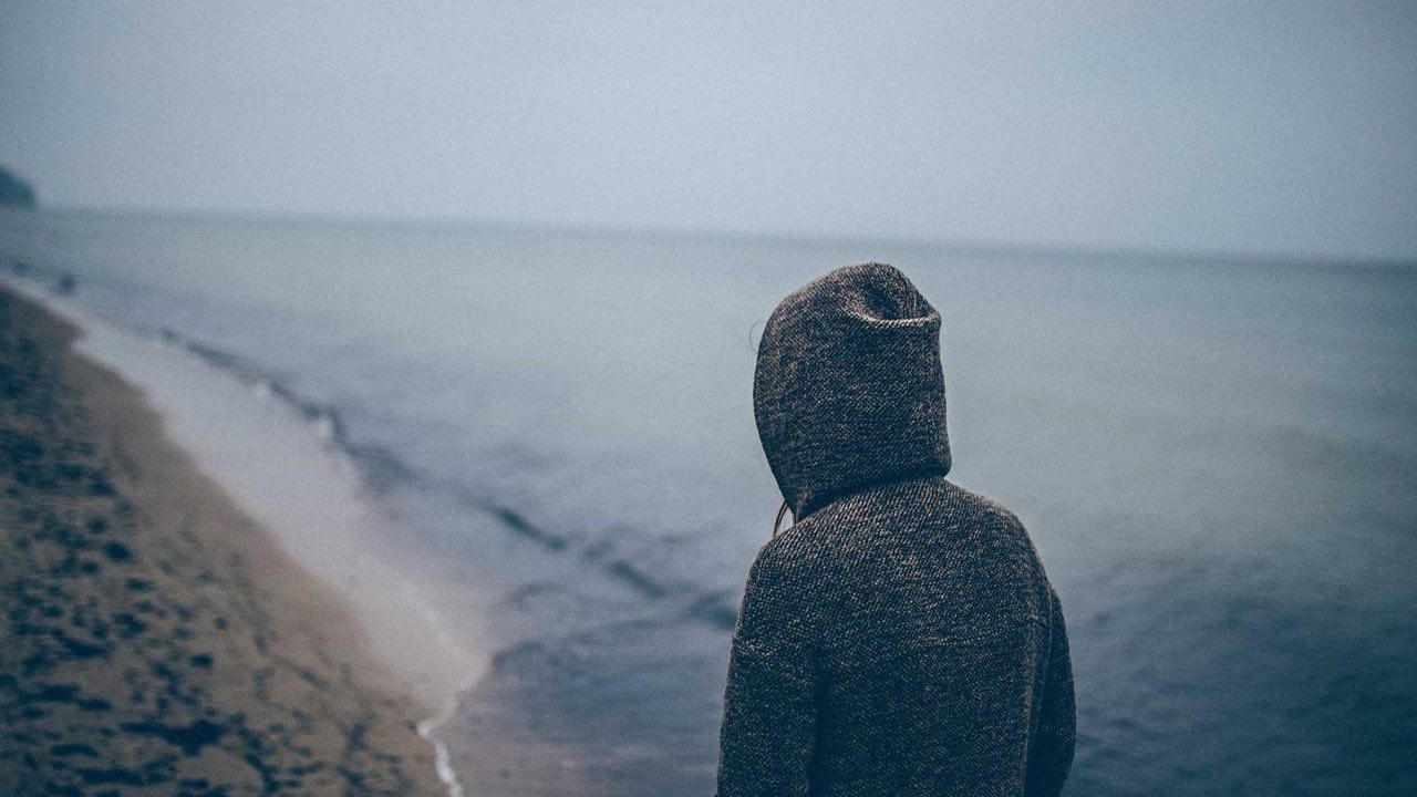 A figure in a hooded coat looking out over a grey shore